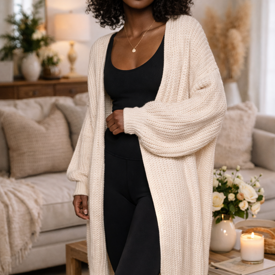 Woman wearing oversized cream knit cardigan with black leggings and tank in a warm, cozy home setting
