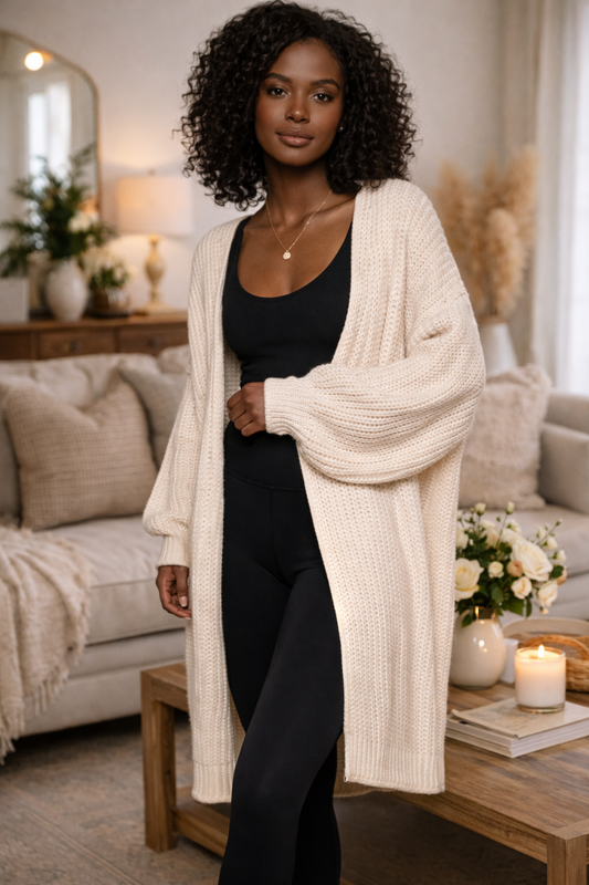 Woman wearing oversized cream knit cardigan with black leggings and tank in a warm, cozy home setting