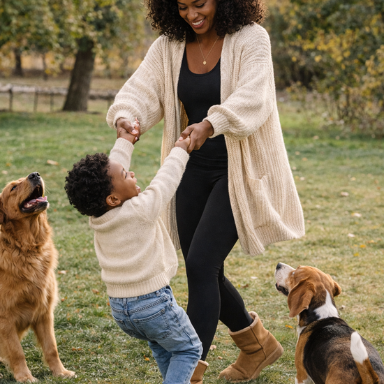 Woman wearing an oversized cream knit cardigan and black leggings playing with a child outdoors in a park with two dogs, capturing a warm and active lifestyle moment