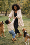 Woman wearing an oversized cream knit cardigan and black leggings playing with a child outdoors in a park with two dogs, capturing a warm and active lifestyle moment