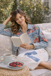 Woman relaxing on an outdoor sofa reading a book while wearing cream patchwork pants and a floral embroidered denim jacket in a soft, sunlit setting
