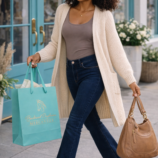 woman wearing an oversized cream knit cardigan with dark bootcut jeans carrying a turquoise shopping bag and brown purse walking outside a boutique with turquoise doors, cozy fall cardigan outfit for women, casual chic boutique style, comfortable layering piece