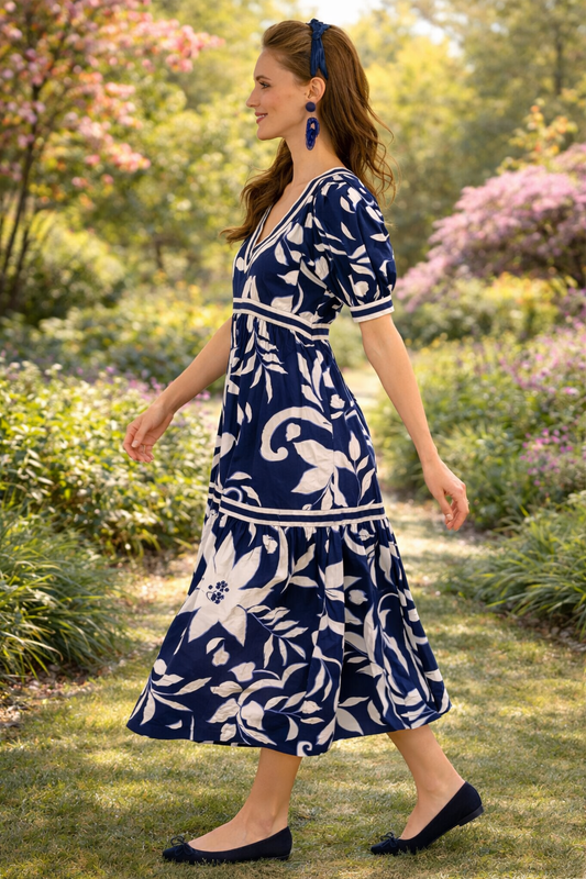 Side view of a woman walking through a sunlit spring garden wearing a navy and white floral midi dress with a flowing silhouette and soft feminine movement