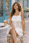 woman sitting at an outdoor café wearing a white tiered mini dress with floral detail and button front, lightweight boho summer dress styled for everyday wear with iced coffee and casual feminine outfit vibes