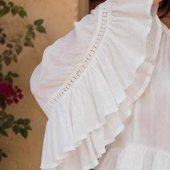 Close-up detail of the Aria Crinkled Ruffle Top featuring dramatic layered bell sleeves, delicate lace trim accents, and lightweight crinkled white fabric in a rustic boho-inspired outdoor setting.