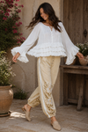 woman twirling in the Aria Crinkled Ruffle Top, a flowy white boho bell sleeve blouse with layered ruffles, paired with embroidered neutral jogger pants in a rustic outdoor courtyard.