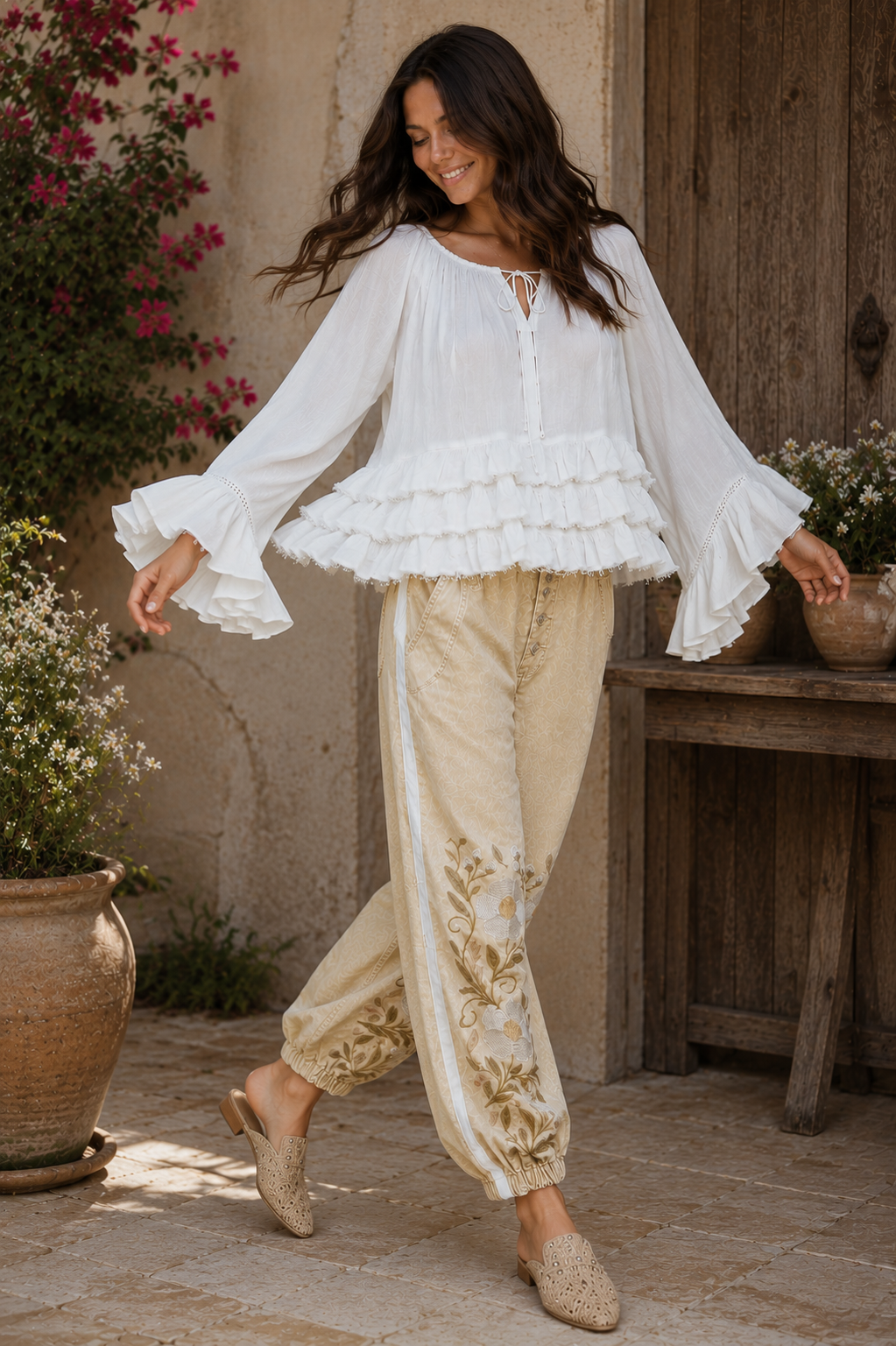 woman twirling in the Aria Crinkled Ruffle Top, a flowy white boho bell sleeve blouse with layered ruffles, paired with embroidered neutral jogger pants in a rustic outdoor courtyard.