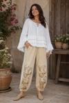 Full-length lifestyle image of a woman wearing the Aria Crinkled Ruffle Top, a white flowy boho blouse with layered ruffles and bell sleeves, paired with embroidered neutral jogger pants in a rustic outdoor courtyard.