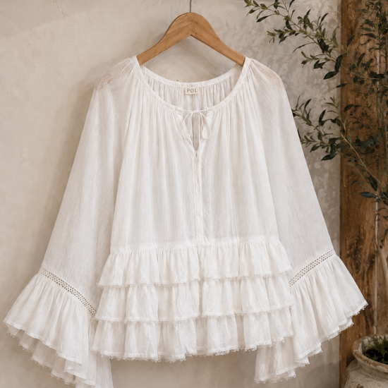 White Aria Crinkled Ruffle Top displayed on a wooden hanger in a rustic boutique-inspired setting, featuring flowy bell sleeves, layered ruffles, lace trim accents, and lightweight crinkled fabric.