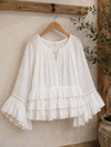 White Aria Crinkled Ruffle Top displayed on a wooden hanger in a rustic boutique-inspired setting, featuring flowy bell sleeves, layered ruffles, lace trim accents, and lightweight crinkled fabric.