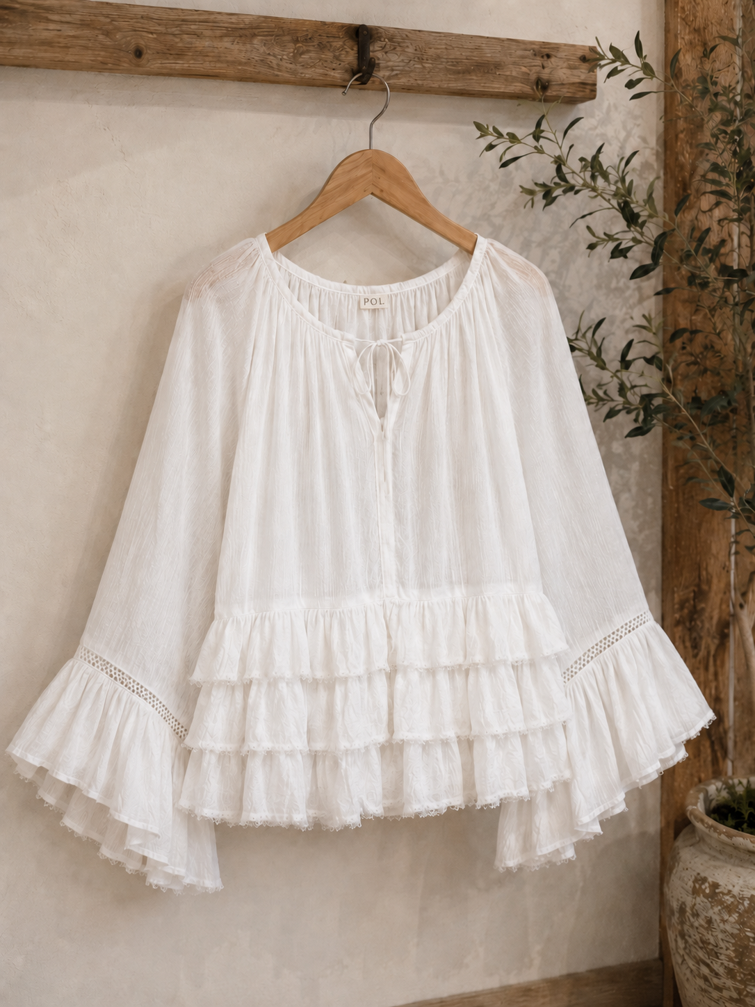 White Aria Crinkled Ruffle Top displayed on a wooden hanger in a rustic boutique-inspired setting, featuring flowy bell sleeves, layered ruffles, lace trim accents, and lightweight crinkled fabric.