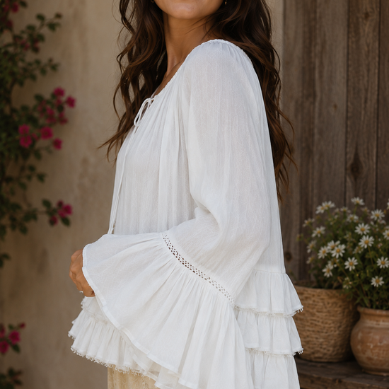 Close-up side view of a brunette woman wearing the Aria Crinkled Ruffle Top, featuring flowy bell sleeves, layered ruffle detailing, and lightweight crinkled white fabric in a rustic outdoor setting.