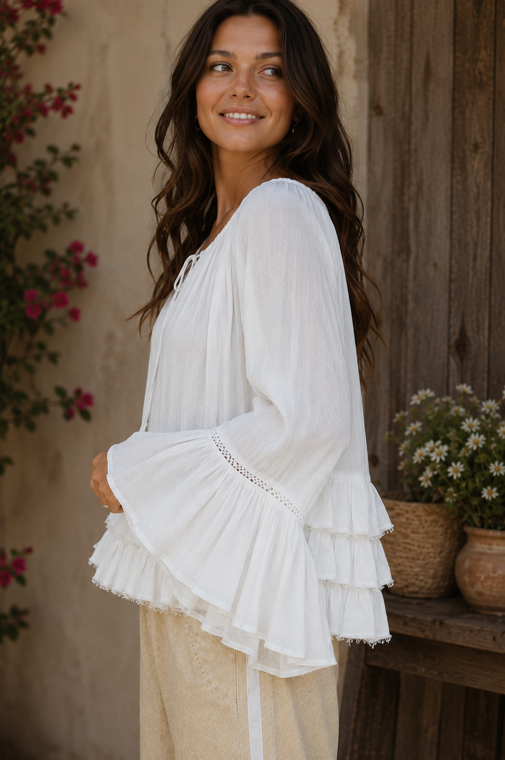 Close-up side view of a brunette woman wearing the Aria Crinkled Ruffle Top, featuring flowy bell sleeves, layered ruffle detailing, and lightweight crinkled white fabric in a rustic outdoor setting.
