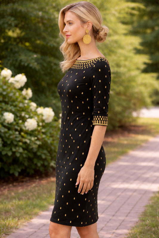 Side view of woman wearing a black sheath dress with gold dotted print and gold neckline detail in an outdoor garden setting.