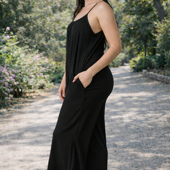 Side view of women's black wide-leg cropped jumpsuit with spaghetti straps styled with flat sandals outdoors