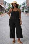 Women's black wide-leg cropped jumpsuit with spaghetti straps styled with wedge sandals and straw hat on a small town street