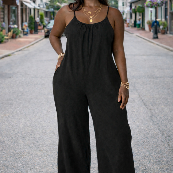 Women's black wide-leg cropped jumpsuit with spaghetti straps styled with straw hat, layered gold jewelry, and wedge sandals on a small town street