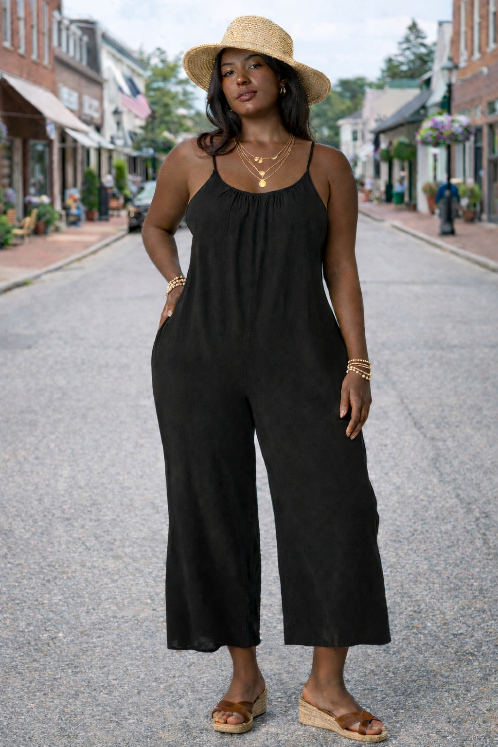 Women's black wide-leg cropped jumpsuit with spaghetti straps styled with straw hat, layered gold jewelry, and wedge sandals on a small town street