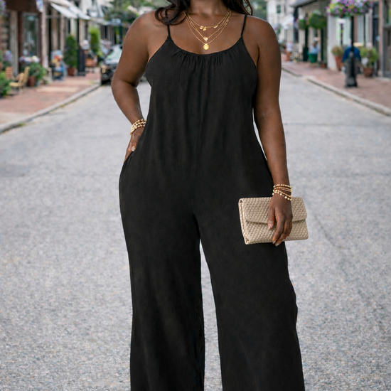 Women's black wide-leg cropped jumpsuit with spaghetti straps styled with wedge sandals, layered gold jewelry, and woven clutch on a small town street