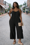 Women's black wide-leg cropped jumpsuit with spaghetti straps styled with wedge sandals, layered gold jewelry, and woven clutch on a small town street