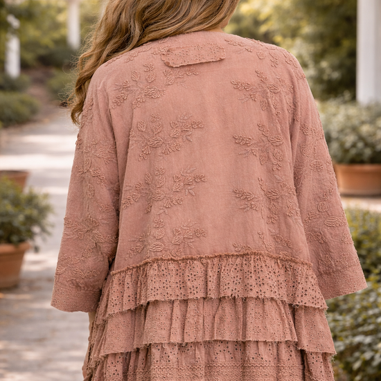 Back view of a  woman wearing a dusty rose BlueVelvet boho embroidered ruffle kimono top with eyelet detailing, styled with sage green pants in an outdoor garden setting