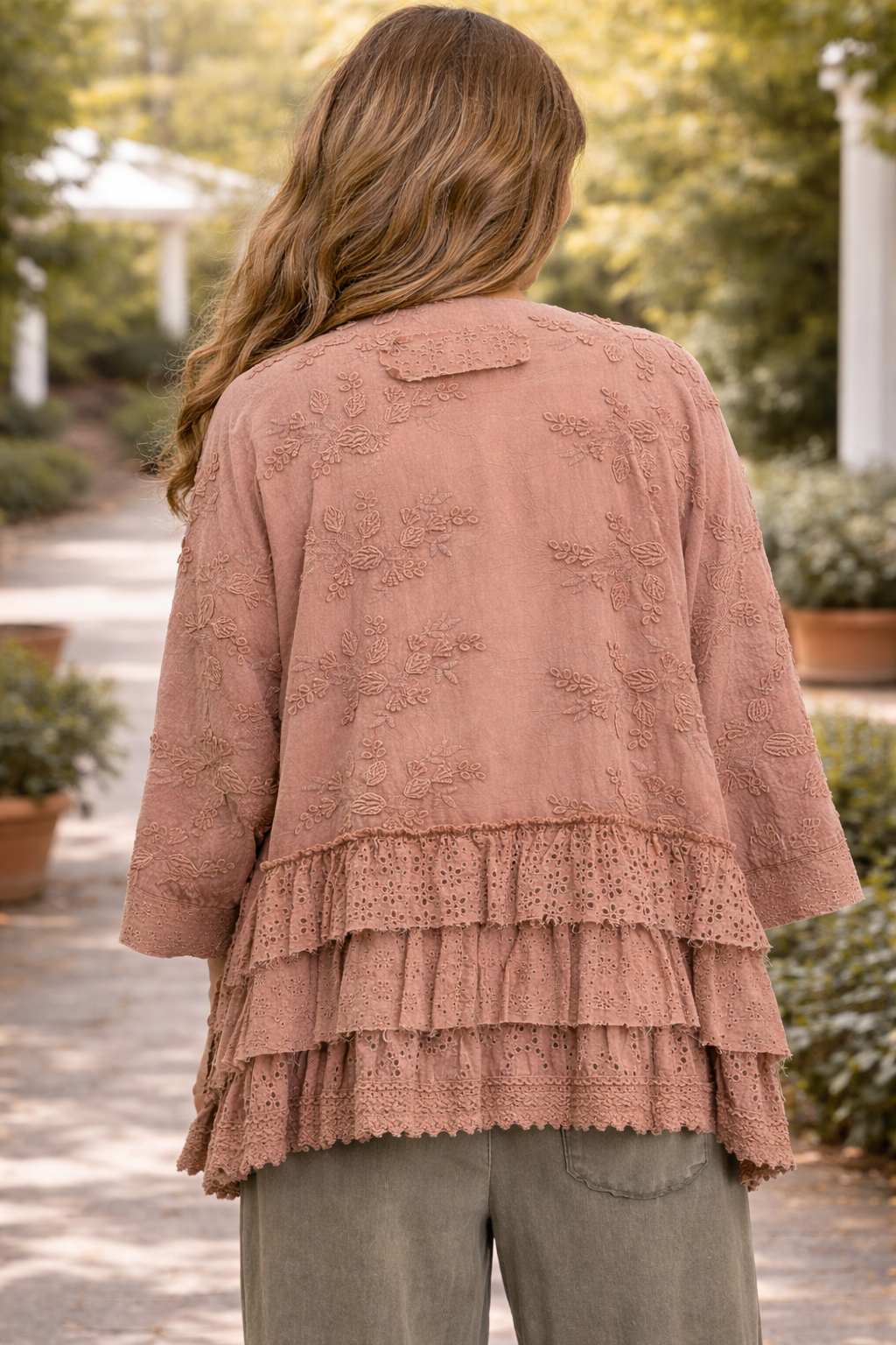 Back view of a  woman wearing a dusty rose BlueVelvet boho embroidered ruffle kimono top with eyelet detailing, styled with sage green pants in an outdoor garden setting
