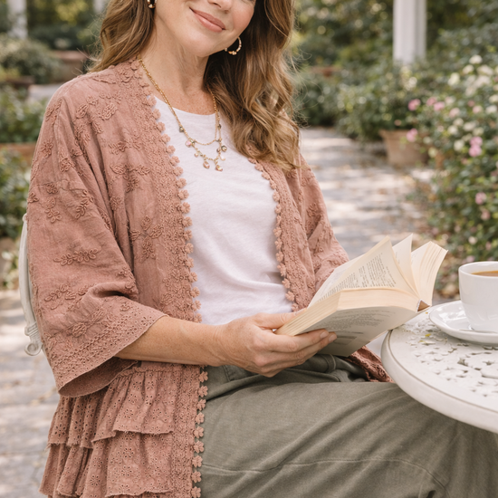 Woman wearing a dusty rose BlueVelvet boho embroidered ruffle kimono with eyelet details, styled with a white tee and sage pants, sitting at an outdoor garden café with a book and coffee