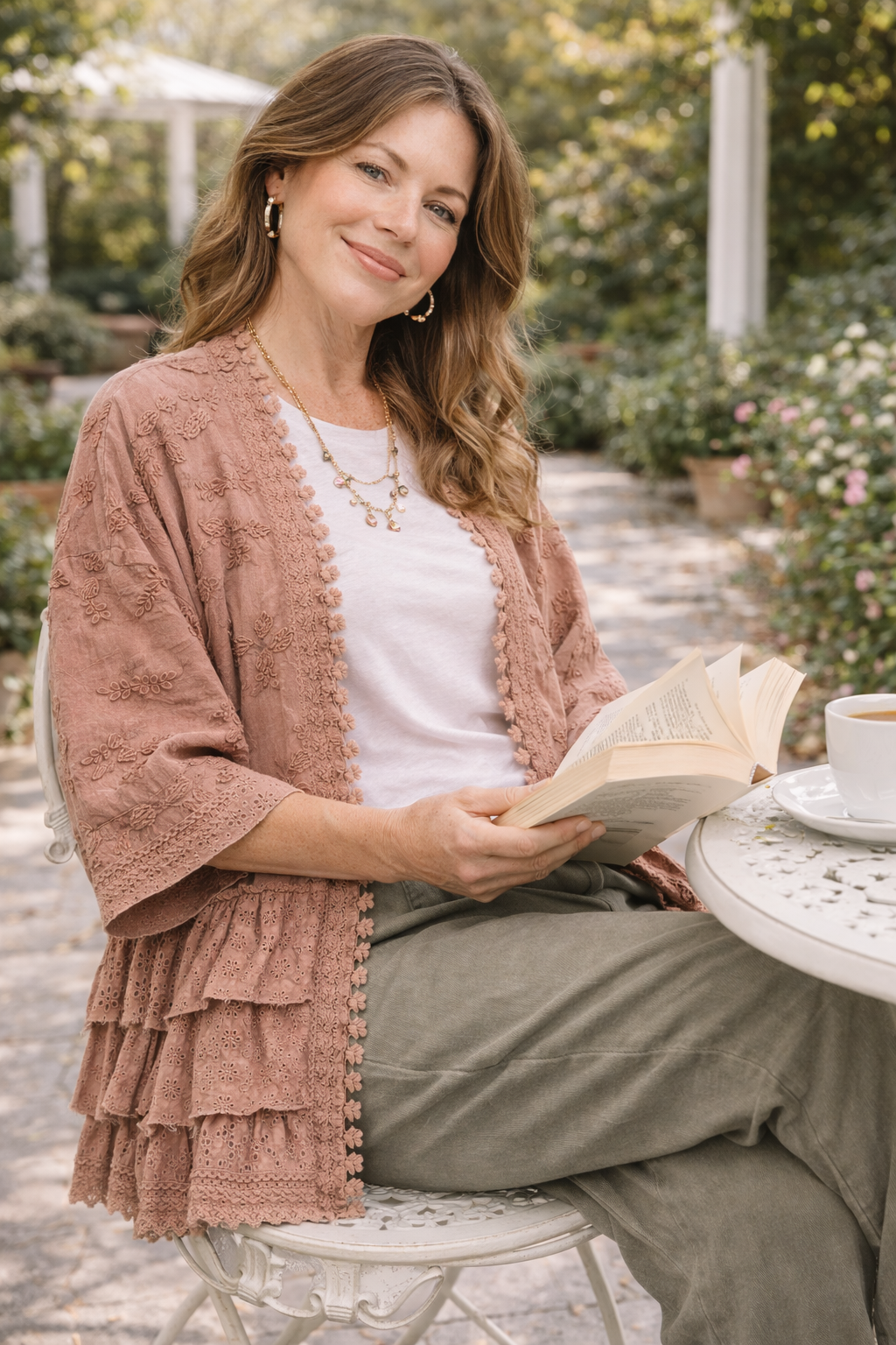 Woman wearing a dusty rose BlueVelvet boho embroidered ruffle kimono with eyelet details, styled with a white tee and sage pants, sitting at an outdoor garden café with a book and coffee