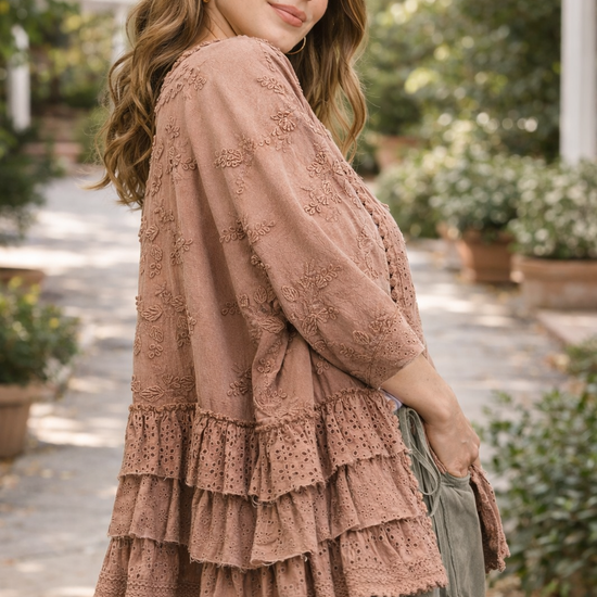woman wearing a dusty rose BlueVelvet boho embroidered ruffle kimono with eyelet details, styled with sage green pants in a soft outdoor garden setting