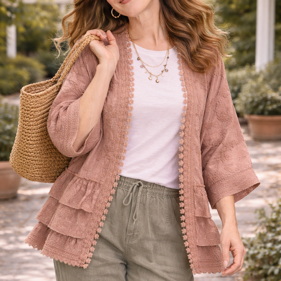 Woman walking through a garden wearing a dusty rose BlueVelvet boho embroidered ruffle kimono with eyelet details, styled with a white tee, sage pants, and a woven tote