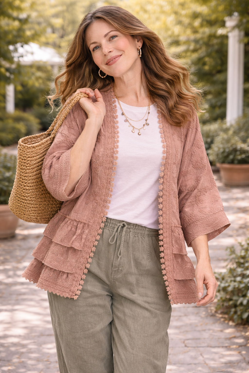 Woman walking through a garden wearing a dusty rose BlueVelvet boho embroidered ruffle kimono with eyelet details, styled with a white tee, sage pants, and a woven tote