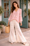 woman wearing blush boho blouse with cream lace wide leg pants standing outside boutique with teal doors in a soft sunlit setting