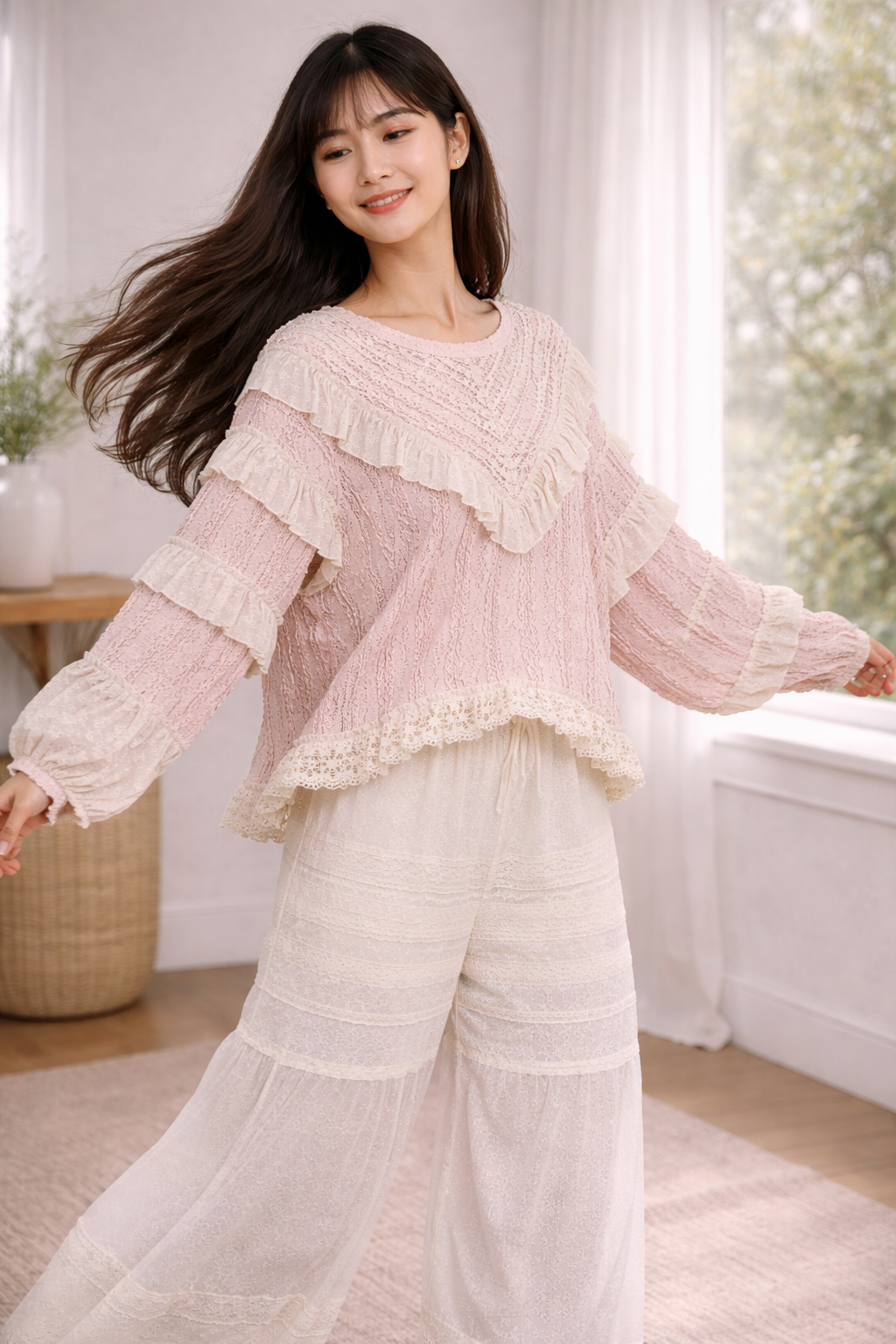 Woman twirling in blush pink boho lace ruffle top and cream wide leg pants in a soft neutral indoor setting, showcasing flowy movement and texture