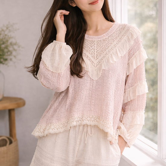 Woman standing by window in blush pink boho lace ruffle top and cream wide leg pants, softly touching her hair in a natural light neutral interior