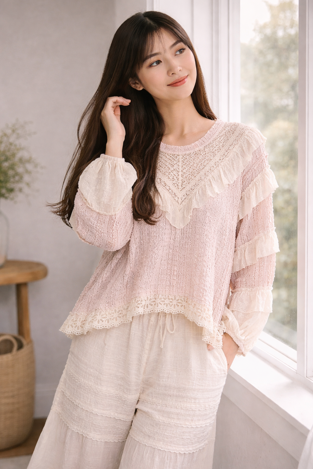 Woman standing by window in blush pink boho lace ruffle top and cream wide leg pants, softly touching her hair in a natural light neutral interior