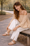Woman sitting on a wooden bench adjusting sandal, wearing a neutral boho off-shoulder blouse and distressed white wide-leg jeans in a relaxed outdoor lifestyle setting