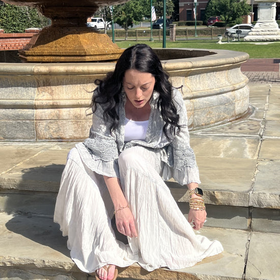 Woman wearing Boho patchwork ruffle lace shrug and Grey boho tiered patchwork pants sitting on steps with a water fountain in the background.