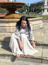 Woman wearing Boho patchwork ruffle lace shrug and Grey boho tiered patchwork pants sitting on steps with a water fountain in the background.