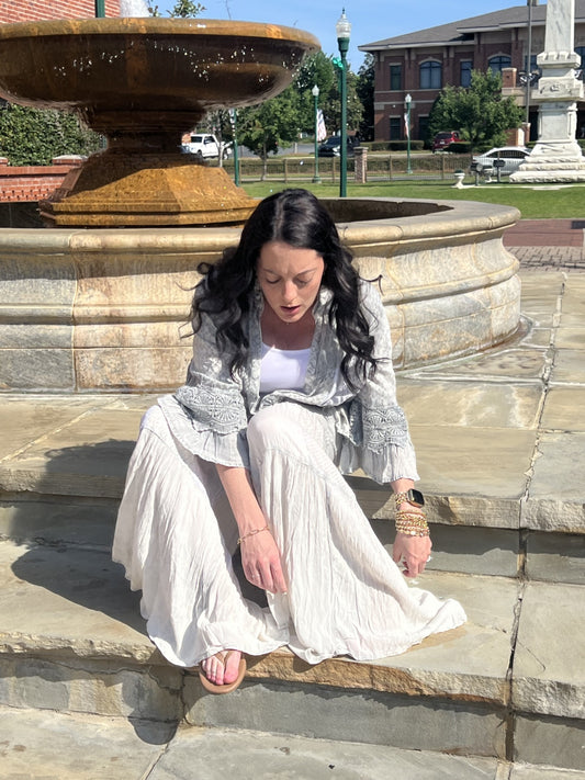 Woman wearing Boho patchwork ruffle lace shrug and Grey boho tiered patchwork pants sitting on steps with a water fountain in the background.