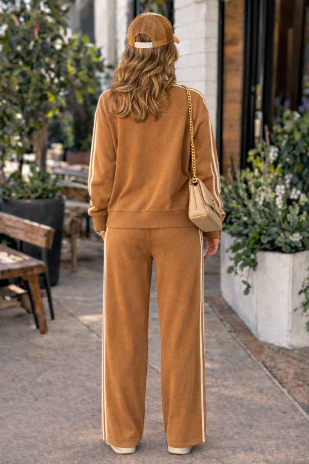 Back view of woman wearing caramel stripe track jacket and wide leg lounge pants athleisure set styled with trucker hat, sneakers, and quilted shoulder bag in casual outdoor setting
