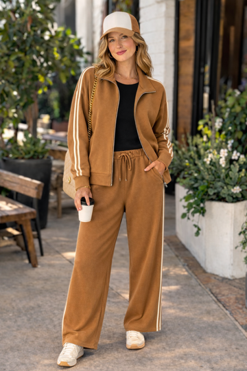 30s woman wearing caramel stripe track jacket and matching wide leg track pants lounge set styled with trucker hat and sneakers in a casual street style outfit