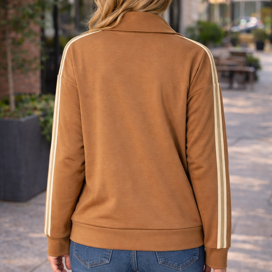 Back view of caramel zip up track jacket with cream stripe sleeves worn by a woman styled with blue jeans in an outdoor setting