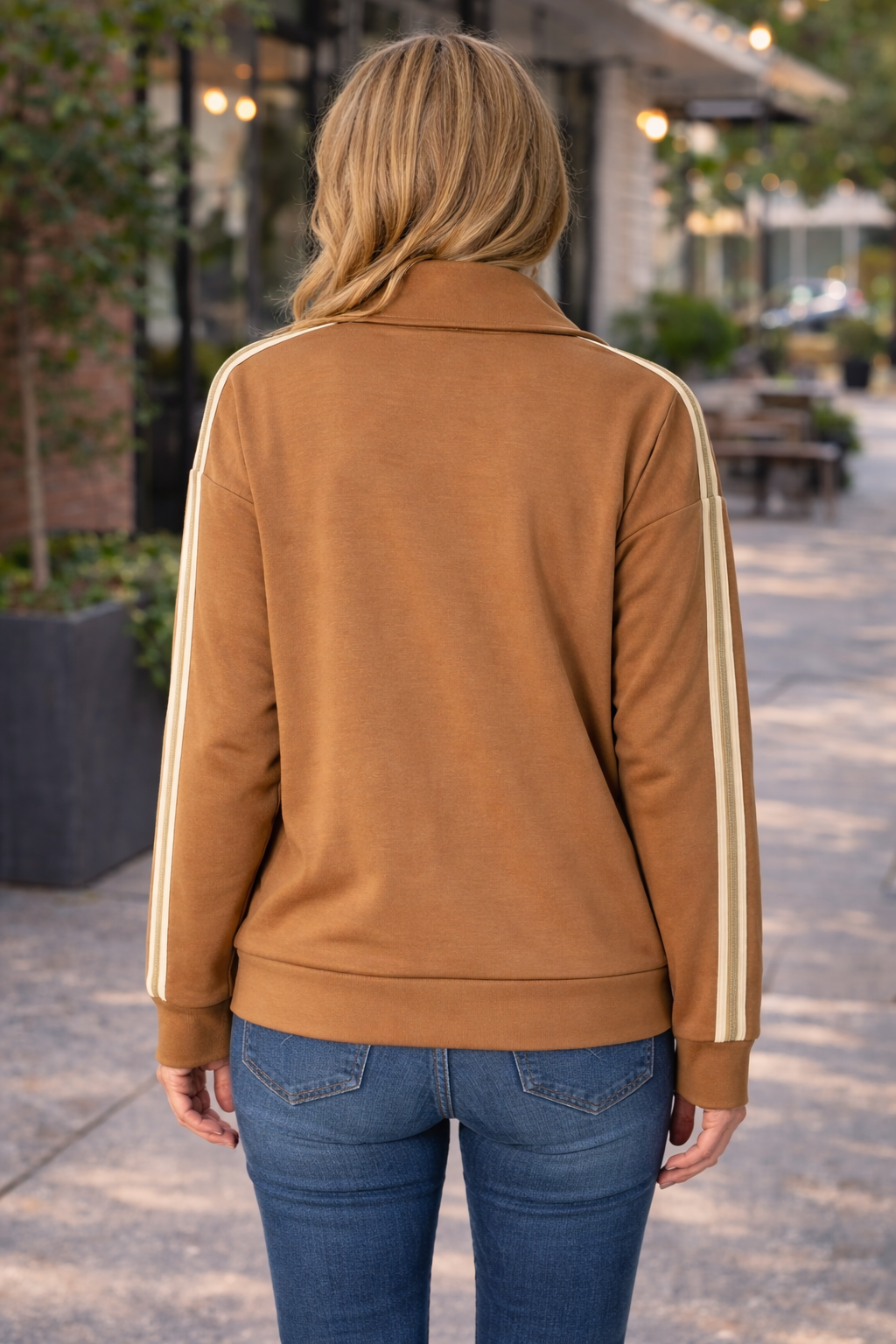 Back view of caramel zip up track jacket with cream stripe sleeves worn by a woman styled with blue jeans in an outdoor setting