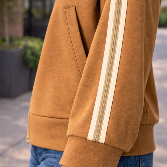 Close up detail of caramel zip up track jacket sleeve with cream stripe trim styled with blue jeans in an outdoor setting