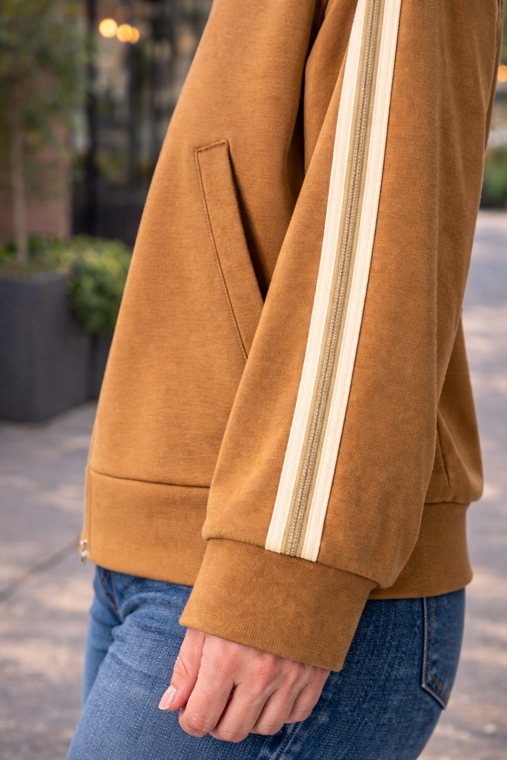 Close up detail of caramel zip up track jacket sleeve with cream stripe trim styled with blue jeans in an outdoor setting