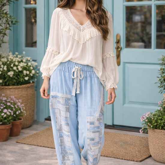 Woman wearing chambray patchwork joggers with cream boho blouse standing outside boutique with turquoise doors and potted flowers