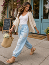 Woman walking in chambray patchwork joggers with white tank and beige cardigan, carrying straw tote outside boutique in relaxed boho style