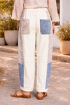 Back view of cream patchwork pants with floral, lace, and denim panels styled with a soft boho blouse and tan leather sandals in a sunlit boutique courtyard