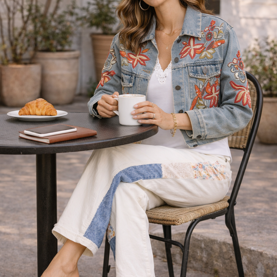 Woman sitting at an outdoor café table wearing cream patchwork pants with floral and denim panels styled with an embroidered denim jacket, white top, and tan sandals while holding a coffee