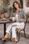 Woman sitting at an outdoor café table wearing cream patchwork pants with floral and denim panels styled with an embroidered denim jacket, white top, and tan sandals while holding a coffee